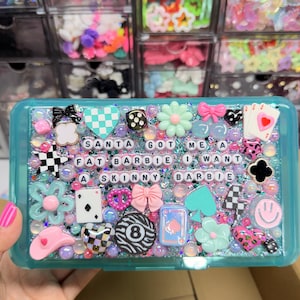May include: A rectangular teal plastic storage box decorated with various charms including flowers, bows, playing cards, dice, and a smiley face. The center features letter beads spelling out the text: SANTA GOT ME A FAT BARBIE I WANT A SKINNY BARBIE.