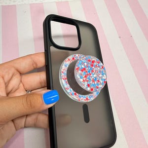 May include: A matte black smartphone case with a translucent back panel and a circular, collapsible phone grip attached to the center. The grip features a clear resin surface embedded with a dense mix of red, white, and blue glitter flakes.