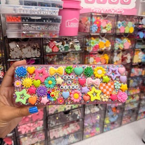 May include: A rectangular storage case decorated with a vibrant assortment of colorful plastic charms including stars, hearts, flowers, bows, and a smiley face. The surface is covered in multi-colored sprinkles and features the text DAY & NIGHT.