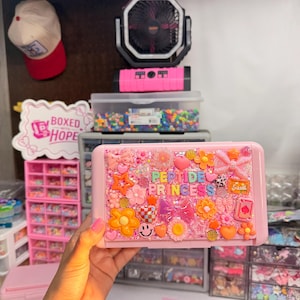 May include: A rectangular pink plastic storage case decorated with various colorful charms including flowers, stars, hearts, bows, and a smiley face. The center features the text PEPTIDE PRINCESS in multi-colored block letters on a glittery background.