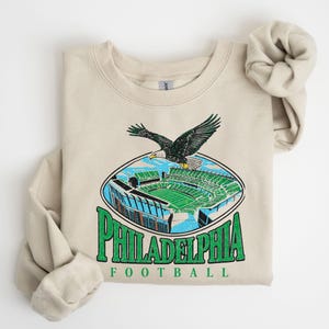 A beige sweatshirt with a graphic design depicting a Philadelphia Eagles football stadium and an eagle in flight.