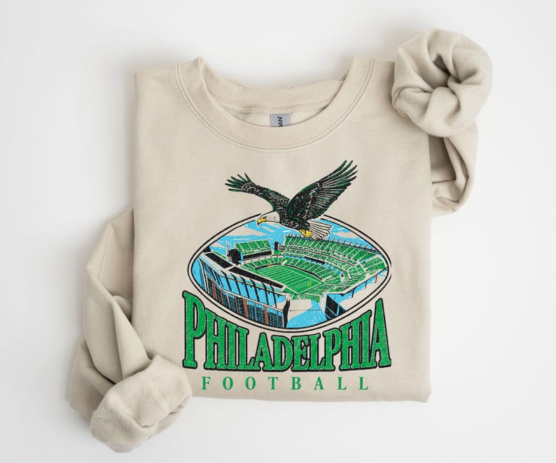 A beige sweatshirt with a graphic design depicting a Philadelphia Eagles football stadium and an eagle in flight.