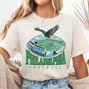 A woman wearing a white t-shirt with a graphic of Philadelphia Eagles football stadium and an eagle in flight.