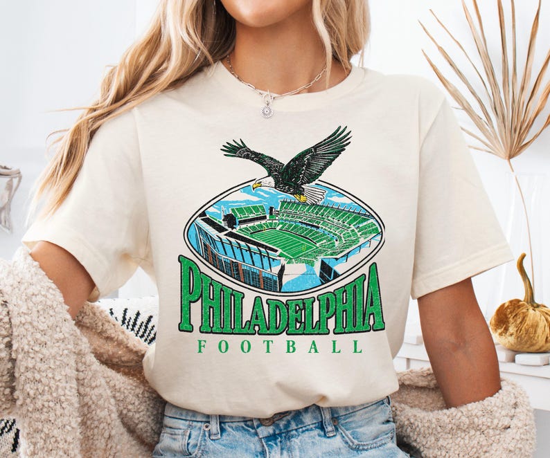 A woman wearing a white t-shirt with a graphic of Philadelphia Eagles football stadium and an eagle in flight.