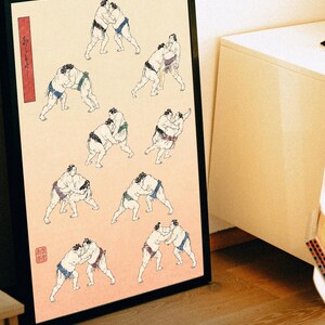 Könnte beinhalten: Ein gerahmter Druck mit einer Reihe von schwarzen Tuscheillustrationen von Sumoringern in verschiedenen Ringkampfpositionen auf hellpfirsichfarbenem Hintergrund. Ein vertikales rotes rechteckiges Etikett mit schwarzem japanischem Text befindet sich auf der linken Seite.