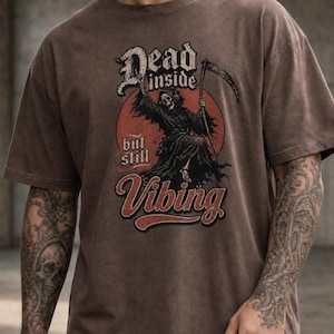 May include: A brown short-sleeve t-shirt featuring a graphic of a skeleton wearing headphones and holding a scythe. The text reads Dead inside but still Vibing. The design is set against a red circular background with a distressed, vintage aesthetic.
