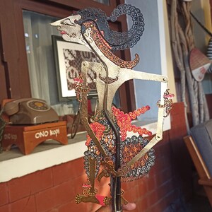 May include: A Javanese shadow puppet, or wayang kulit, featuring an intricate, perforated design in gold, black, and red. The figure has a pointed nose, elaborate headpiece, and articulated arms with rods, showcasing detailed patterns throughout the body.