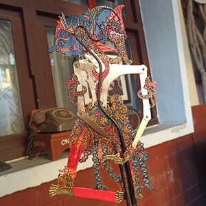 Könnte beinhalten: Eine javanische Schattenfigur mit einem blau bemalten Gesicht und komplexen Mustern in Rot, Gold und Schwarz. Die Figur verfügt über bewegliche Gliedmaßen, die mit Metallstiften verbunden sind, und ist an einem schwarzen Stab befestigt.