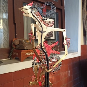 Pode incluir: Uma marioneta de sombras wayang kulit javanesa com uma figura pintada a ouro e padrões de filigrana pretos e vermelhos intrincados. A marioneta tem braços articulados com pinos de metal nas juntas e uma haste preta central para movimento.