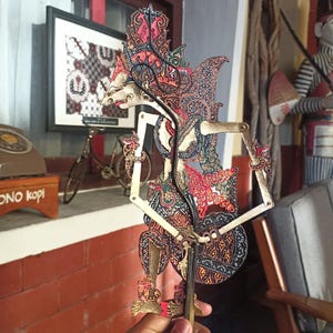Könnte beinhalten: Eine traditionelle javanische Wayang-Kulit-Schattenfigur mit komplexen Ausschnittmustern in Schwarz, Gold und Rot. Die Figur hat eine spitze Kopfbedeckung, bewegliche Arme mit dünnen Stäben und detaillierte Filigranarbeiten am ganzen Körper.