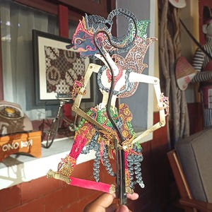 May include: A traditional Javanese wayang kulit shadow puppet featuring an intricate, hand-painted figure with a dark blue face, ornate gold-toned filigree details, and vibrant red, green, and black patterns on the clothing, mounted on a black rod.