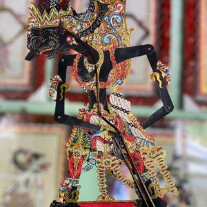 Könnte beinhalten: Eine traditionelle javanische Schattenfigur, bekannt als Wayang Kulit, mit detaillierten Bemalungen in Schwarz, Gold, Rot und Blau. Die bewegliche Figur zeigt einen stilisierten Kopfschmuck, verzierte Kleidungsmuster und einen zentralen Steuerstab.