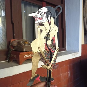 Puede incluir: Un títere de sombra javanés, o wayang kulit, que presenta una figura estilizada con cara blanca, cabello negro y cuerpo de color dorado. El títere viste un pareo estampado con detalles en rojo, azul y negro y está unido a un mango central.