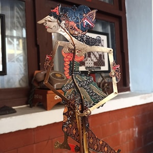 Puede incluir: Una marioneta de sombras wayang kulit javanesa con un perfil alargado y detalles intrincados pintados a mano en negro, oro, rojo y verde. La figura tiene brazos articulados con varillas metálicas finas para el movimiento y una prenda estampada.