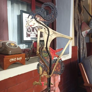 May include: A Javanese shadow puppet, or wayang kulit, featuring intricate black and gold filigree patterns. The figure has a pointed nose, a curved headdress, and articulated limbs connected by metal pins, with red and brown fabric accents.