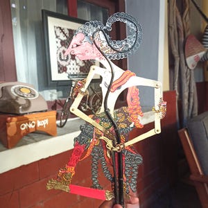 Könnte beinhalten: Eine javanische Schattenfigur, oder Wayang Kulit, mit komplizierten Ausschnitten und handgemalten Mustern in Rosa, Schwarz, Gold und Rot. Die Figur hat einen langen, geschwungenen Kopfschmuck, bewegliche Arme mit dünnen Stäben und einen Sockel.