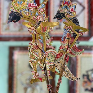 Puede incluir: Dos marionetas de sombra intrincadas con rostros negros y elaborados tocados coloridos con patrones dorados, rojos, azules y blancos. Cada marioneta está unida a una varilla de madera vertical con brazos articulados de color dorado.