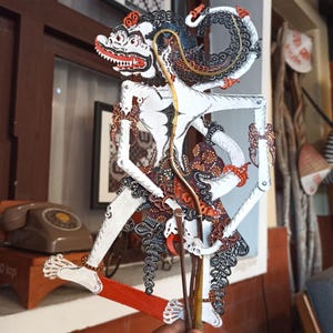 Puede incluir: Una marioneta de sombras wayang kulit javanesa que representa un personaje de piel blanca con detalles pintados en negro y rojo, extremidades articuladas y una varilla de control de bambú, con patrones tradicionales y ornamentados.