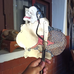 Puede incluir: Una marioneta de sombras javanesa tradicional, o wayang kulit, que presenta una figura estilizada con cara blanca, cabello negro y lengua roja. El títere tiene secciones corporales doradas, una falda con estampado batik y una varilla negra.