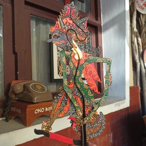 以下が含まれることがあります： 手描きの緑色のボディ、赤と金のアクセント、精巧なフィリグリーの切り抜きが施された、伝統的なジャワのワヤン・クリット影絵人形。人形は水平なハンドルが付いた垂直のロッドに取り付けられています。