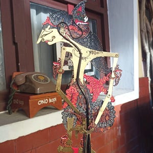Pode incluir: Uma marioneta de sombras wayang kulit javanesa com uma figura intrincada pintada à mão, nariz pontiagudo, um elaborado toucado preto e vermelho e braços articulados. A marioneta é feita de couro com recortes detalhados e detalhes dourados.