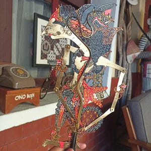 Puede incluir: Una marioneta de sombra javanesa tradicional, o wayang kulit, con intrincados detalles recortados y acentos pintados a mano en oro, negro y rojo. La figura tiene brazos articulados con varillas delgadas para el movimiento y un perfil estilizado.