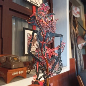 May include: A traditional Javanese shadow puppet, known as a wayang kulit, featuring intricate black, red, and gold filigree patterns. The puppet has a tall, pointed headdress, articulated limbs with thin rods, and a central handle for manipulation.