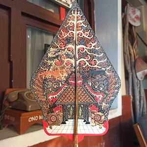 Op de afbeelding: Een traditionele Indonesische wayang kulit schaduwpop, bekend als gunungan of kayon, met ingewikkelde zwarte, rode en gouden uitgesneden patronen van een centrale boom, mythische wezens en een tempelstructuur op een bladvormig frame.