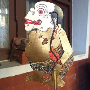 Op de afbeelding: Een Javaanse schaduwpop met een wit gezicht, rode ogen en lippen, een grote goudkleurige romp en beweegbare armen. De figuur draagt een kledingstuk met een patroon in rood, zwart en wit en een gouden ketting.