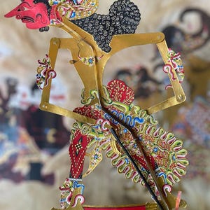 Könnte beinhalten: Eine Wayang-Kulit-Schattenfigur mit einem rot bemalten Gesicht und detaillierten Verzierungen in Gold, Schwarz und bunten Farben. Die Figur verfügt über bewegliche Arme mit dünnen Stäben, einen hohen Kopfschmuck und ein gemustertes Gewand.