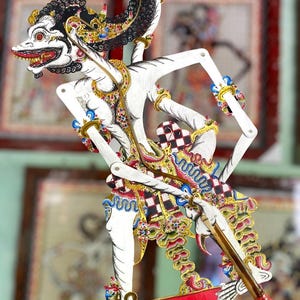 Può includere: Una tradizionale marionetta d'ombra giavanese, o wayang kulit, che raffigura un personaggio dalla pelle bianca con intricati dettagli dipinti in nero, oro, rosso e blu. La figura ha arti articolati fissati a sottili aste di legno.