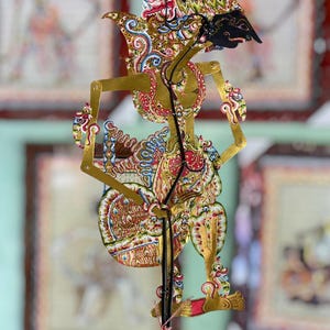 Op de afbeelding: Een traditionele Javaanse schaduwpop, bekend als wayang kulit, met ingewikkelde goudkleurige, rode en zwarte geschilderde details op een platte, geperforeerde lederen figuur. De pop is bevestigd aan een dunne zwarte verticale staaf.