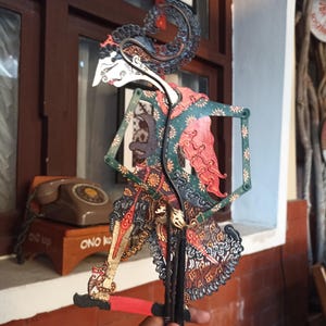 Può includere: Un burattino d'ombra giavanese, o wayang kulit, caratterizzato da intricati motivi ritagliati e dettagli dipinti nelle tonalità del verde, rosso, nero e oro. La figura presenta un profilo stilizzato, un alto copricapo ornato e arti articolati.