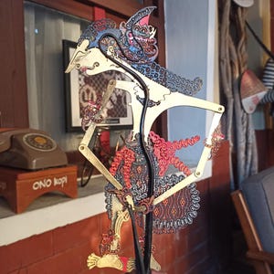 Peut inclure: Une marionnette d'ombre plate et articulée présentant des motifs découpés complexes dans des tons de bleu, rouge et or. La silhouette possède un profil stylisé, des membres allongés et une jupe décorative, fixée sur une fine tige noire.