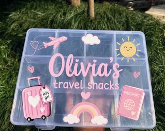 May include: A clear plastic storage container with twelve compartments, featuring pink decals of a suitcase, a passport, a rainbow, a sun, a plane, and hearts. The text "Olivia's travel snacks" is printed in pink script and sans-serif font.