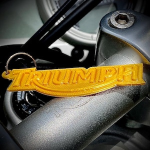May include: A gold-coloured keychain featuring the word TRIUMPH in a raised, textured font. The keychain is attached to a circular metal ring and is positioned against a metallic motorcycle handlebar component with a visible bolt head.