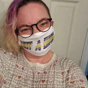 May include: A person wearing a white face mask with the text "Not a Girl" in yellow and purple. The mask has a black and white striped pattern. The person is wearing a white shirt with a pattern of hearts, stars, and skulls.