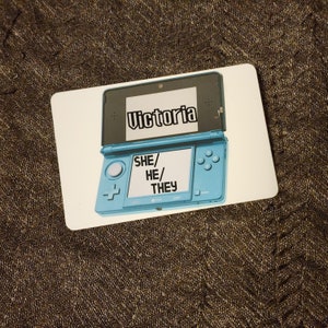 Puede incluir: Una consola de juegos port&aacute;til Nintendo 3DS azul y blanca con el nombre "Victoria" en la pantalla superior y las palabras "SHE/HE/THEY" en la pantalla inferior.