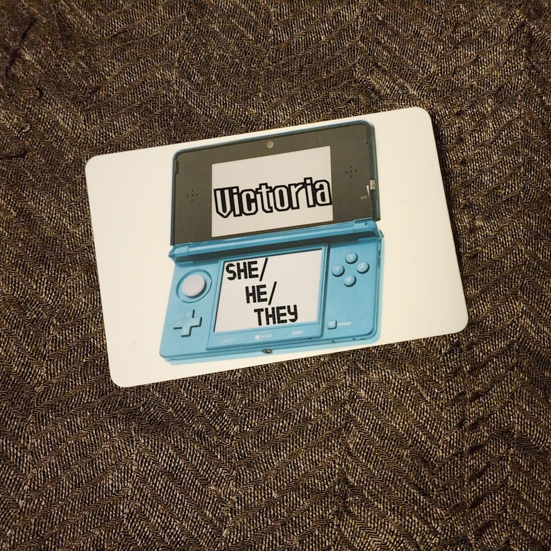 Puede incluir: Una consola de juegos port&aacute;til Nintendo 3DS azul y blanca con el nombre "Victoria" en la pantalla superior y las palabras "SHE/HE/THEY" en la pantalla inferior.