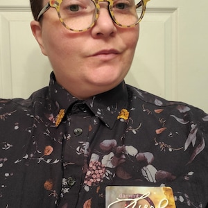 Pu&ograve; includere: Una persona che indossa una camicia nera a fiori con bottoni e un cartellino con su scritto "dammit just xey/xem/xeir's gender neutral language please".