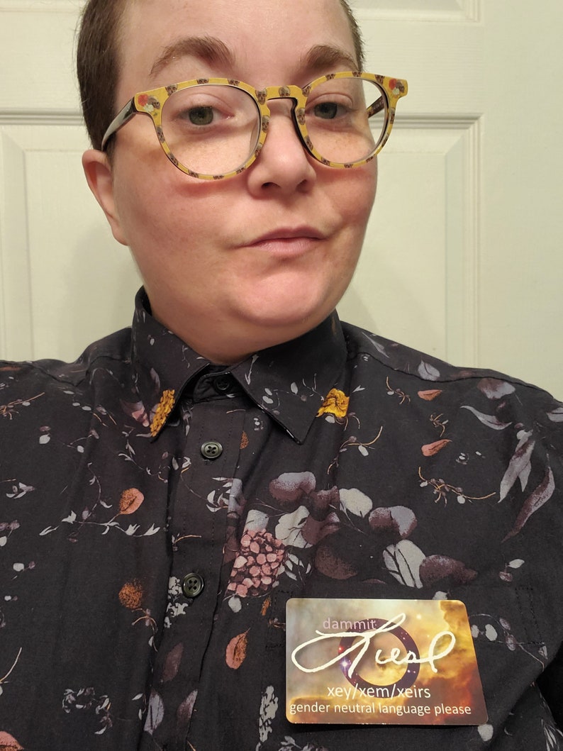 Pu&ograve; includere: Una persona che indossa una camicia nera a fiori con bottoni e un cartellino con su scritto "dammit just xey/xem/xeir's gender neutral language please".
