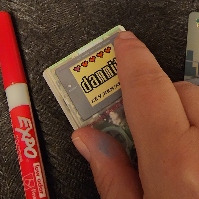 Puede incluir: Un primer plano de un cartucho de juego de Game Boy Color con una pegatina que dice "damnit" y tiene un dise&ntilde;o de coraz&oacute;n pixelado. El cartucho es gris y tiene un compartimento para bater&iacute;as.