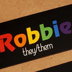 May include: Black name tag with the name "Robbie" in rainbow letters and the text "they/them" in white letters.