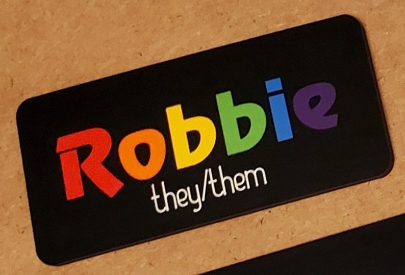 May include: Black name tag with the name "Robbie" in rainbow letters and the text "they/them" in white letters.