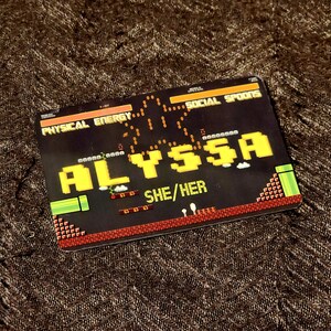 Puede incluir: Una tarjeta negra y roja con tem&aacute;tica de videojuego. La tarjeta tiene un nombre pixelado amarillo "ALYSSA" y el texto "SHE/HER" en blanco. La tarjeta tiene un gr&aacute;fico de un personaje de videojuego saltando sobre un tubo.