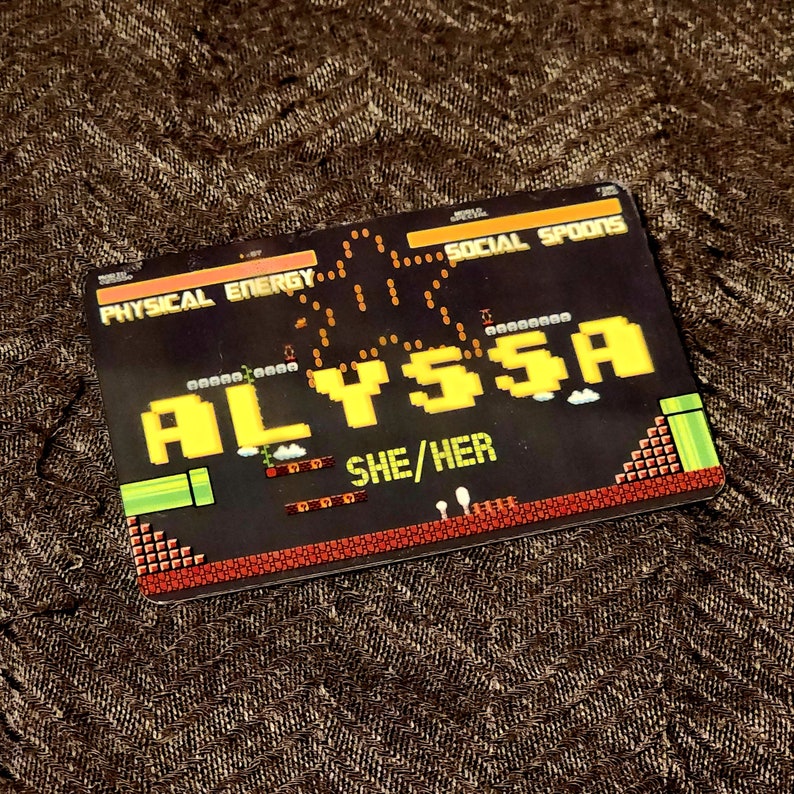 Puede incluir: Una tarjeta negra y roja con tem&aacute;tica de videojuego. La tarjeta tiene un nombre pixelado amarillo "ALYSSA" y el texto "SHE/HER" en blanco. La tarjeta tiene un gr&aacute;fico de un personaje de videojuego saltando sobre un tubo.