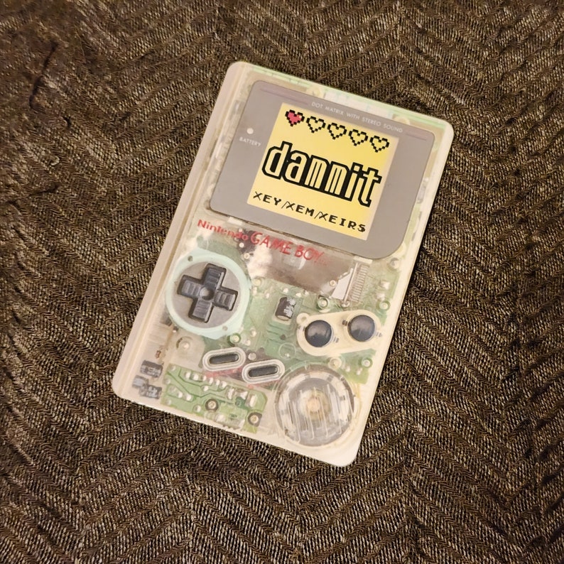 Puede incluir: Una Game Boy transparente con un bot&oacute;n verde y una pegatina que dice "dammit" con un gr&aacute;fico de coraz&oacute;n pixelado. La pegatina tambi&eacute;n dice "XEY/XEM/XEIRS" debajo. La Game Boy est&aacute; sobre una superficie marr&oacute;n texturizada.