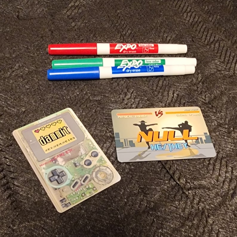 Puede incluir: Un cartucho de juego de Game Boy Color de pl&aacute;stico transparente con una etiqueta amarilla que dice "dammit" y "XEY/XEM/XEERS". El cartucho est&aacute; sobre una superficie marr&oacute;n con tres marcadores de borrado en seco.