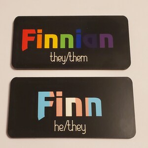 May include: Two black rectangular name tags with white text. The top tag says "Finnian they/them" in rainbow colours. The bottom tag says "Finn he/they" in light blue, pink, and white.