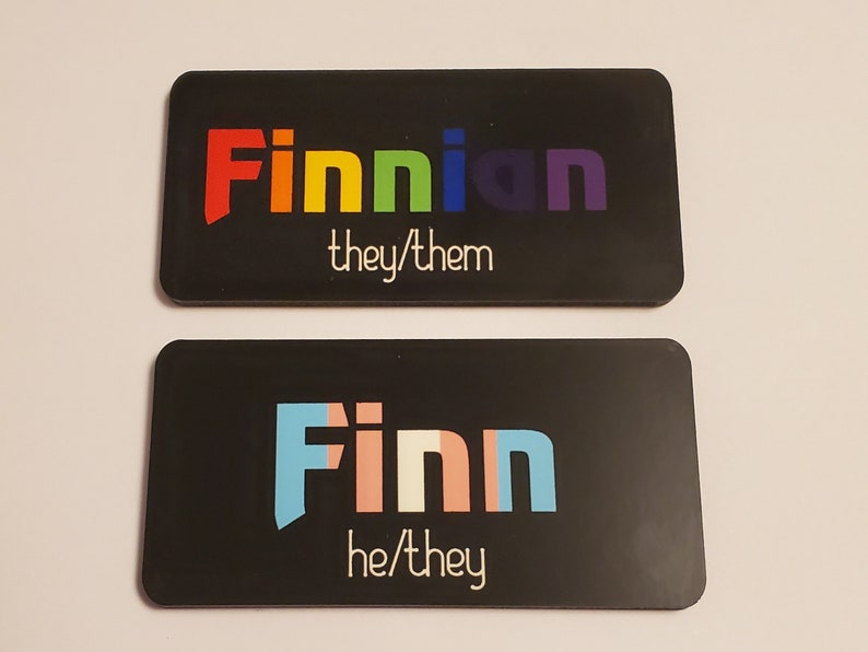 May include: Two black rectangular name tags with white text. The top tag says "Finnian they/them" in rainbow colours. The bottom tag says "Finn he/they" in light blue, pink, and white.